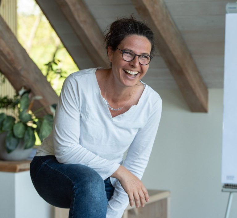 Der Coach in Action Tanja in einem hellen Raum, steht lässig mit Pflanzen im Hintergrund.